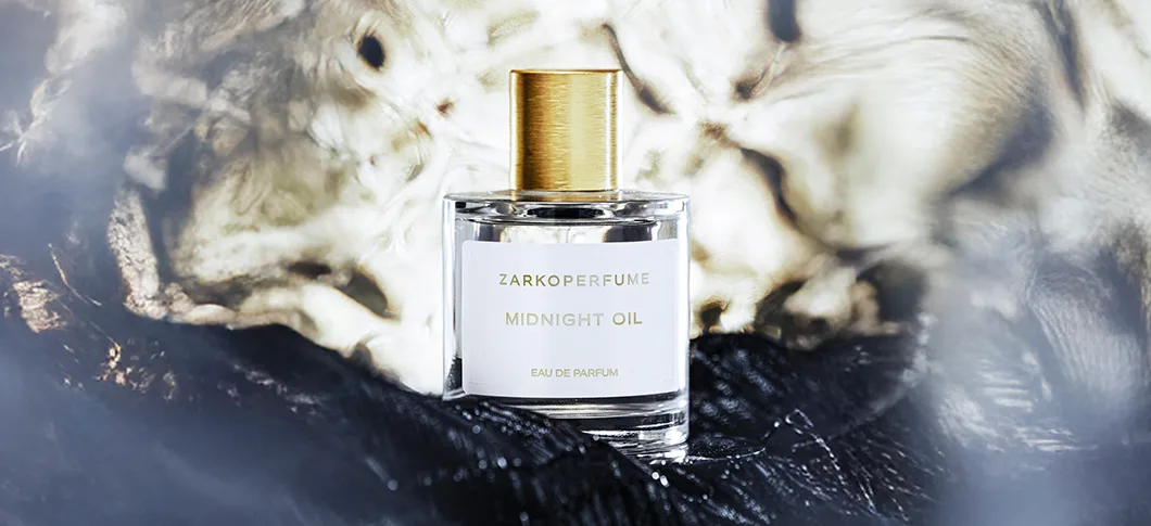 Her ses et billede af en parfumeflakon fra Zarkoperfume placeret centralt mod en mørk og abstrakt baggrund. På flakonen står teksten “ZARKOPERFUME MIDNIGHT OIL EAU DE PARFUM”. Flakonen er i klart glas med guldfarvet låg.