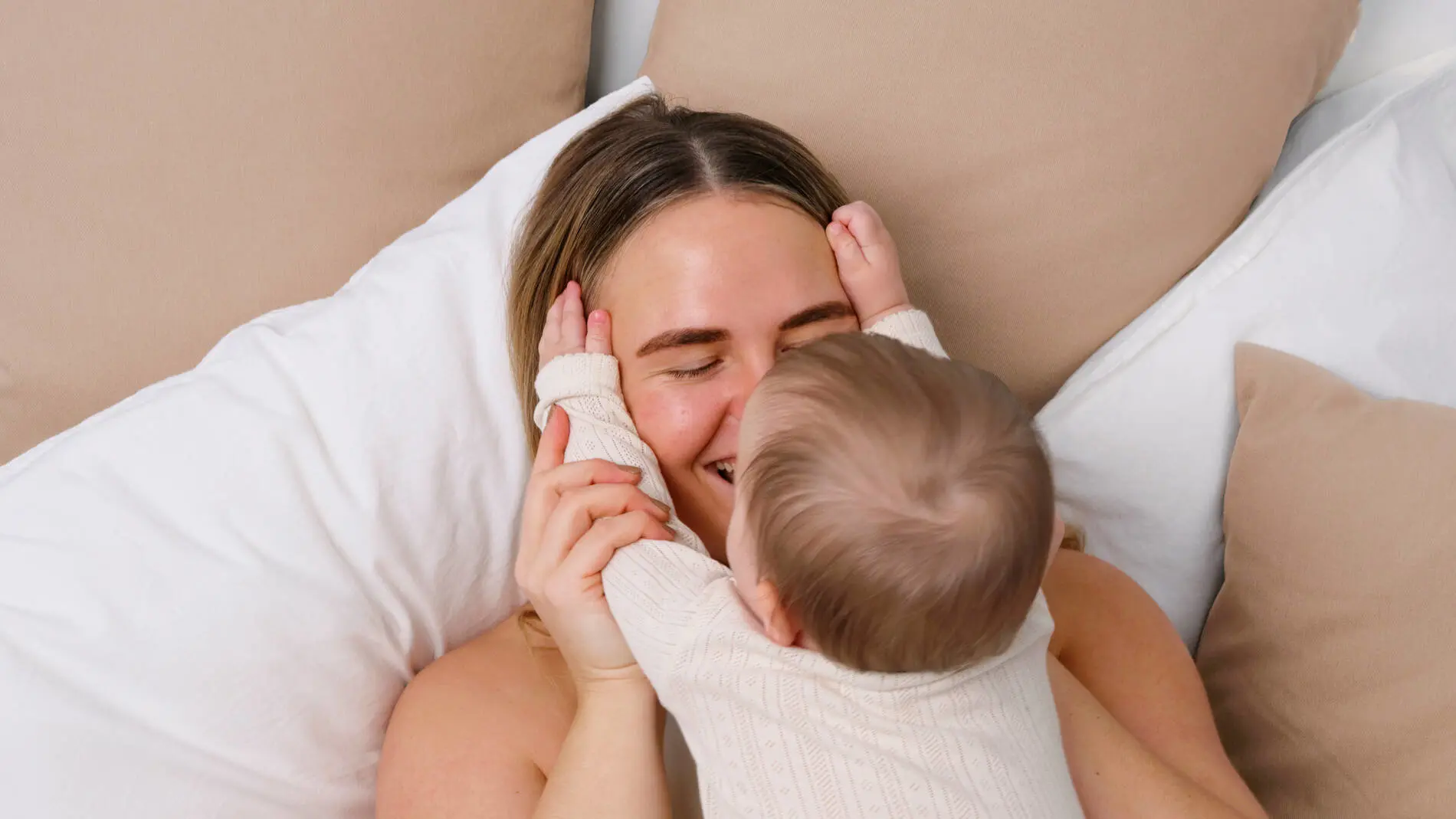 En voksen og et lille barn ligger tæt sammen i en seng omgivet af store hvide og beige puder. Barnet holder sine hænder på den voksnes ansigt og læner sig ind til et kærligt kram, mens den voksne smiler med lukkede øjne. Begge har lyst tøj på, og billedet har en varm, rolig og nærværende stemning.