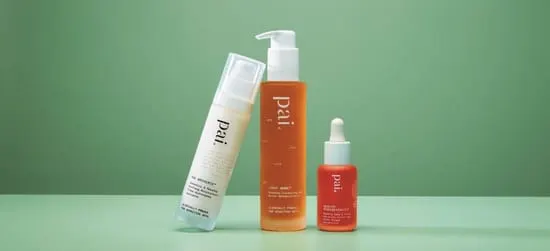 Tre hudplejeprodukter fra Pai står på en lysegrøn flade: en hvid sprayflaske, en høj orange renseflaske med pumpe og en mindre orange serumflaske med pipette.