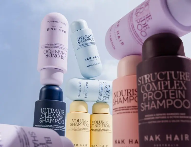 Her ses et billede med hårplejeprodukter fra NAK Hair placeret mod en baggrund med blå himmel. Produkterne er stablet i forskellige højder og omfatter blandt andet “Hydrate Shampoo”, “Hydrate Conditioner”, “Volume Shampoo”, “Volume Conditioner”, “Blonde Shampoo”, “Nourish Shampoo”, “Ultimate Cleanse Shampoo” og “Structure Complex Protein Shampoo”.
