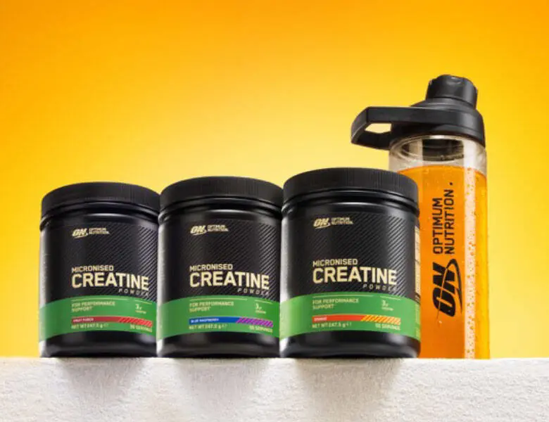 Billedet viser tre sorte bøtter med Optimum Nutrition Micronised Creatine Powder (flavoured creatine monohydrat) med grønne etiketter og forskellige smagsindikatorer, placeret side om side på en lys overflade. Til højre står en klar træningsshaker med Optimum Nutrition-logo fyldt med orange væske. Baggrunden er en jævn gul-orange gradient, der fremhæver produkterne.