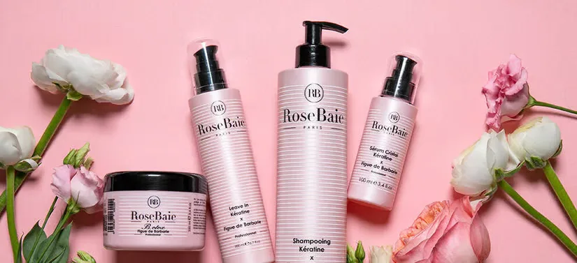 Her ses fire Rosebaie Paris produkter, liggende blandt blomster på en lyserød baggrund.