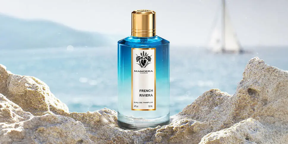 Her ses et billede af en parfumeflakon fra Mancera placeret på klipper ved havet. På flakonen står teksten “MANCERA FRENCH RIVIERA EAU DE PARFUM”. Flakonen er i blå nuancer med guldfarvet låg.
