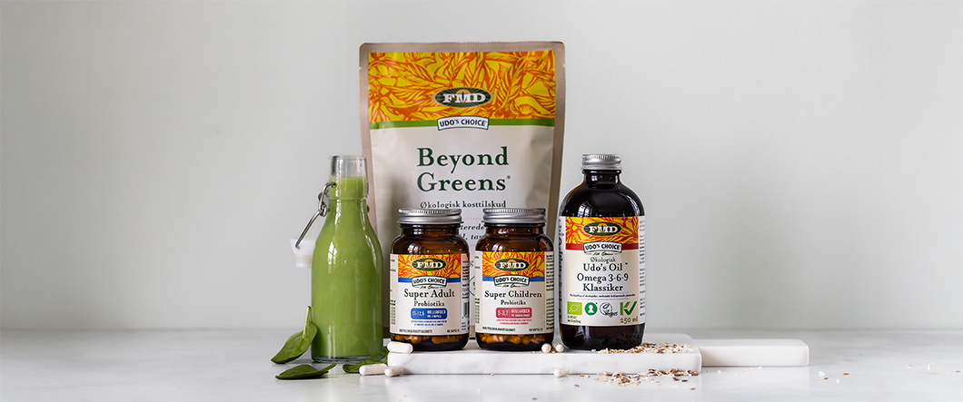 Udo’s Choice kosttilskud står samlet på en lys overflade foran en neutral grå baggrund, herunder Beyond Greens, probiotika til voksne og børn samt omega 3-6-9 olie, sammen med en flaske grøn smoothie, blade, kapsler og frø.