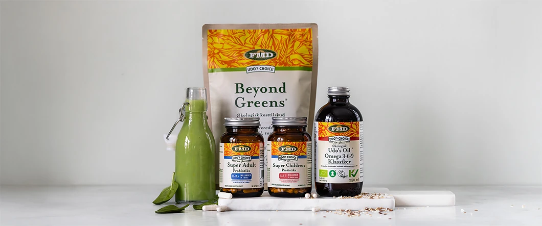 Udo’s Choice kosttilskud står samlet på en lys overflade foran en neutral grå baggrund, herunder Beyond Greens, probiotika til voksne og børn samt omega 3-6-9 olie, sammen med en flaske grøn smoothie, blade, kapsler og frø.