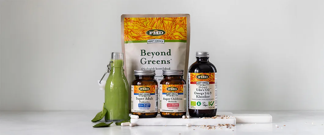 Udo’s Choice kosttilskud står samlet på en lys overflade foran en neutral grå baggrund, herunder Beyond Greens, probiotika til voksne og børn samt omega 3-6-9 olie, sammen med en flaske grøn smoothie, blade, kapsler og frø.