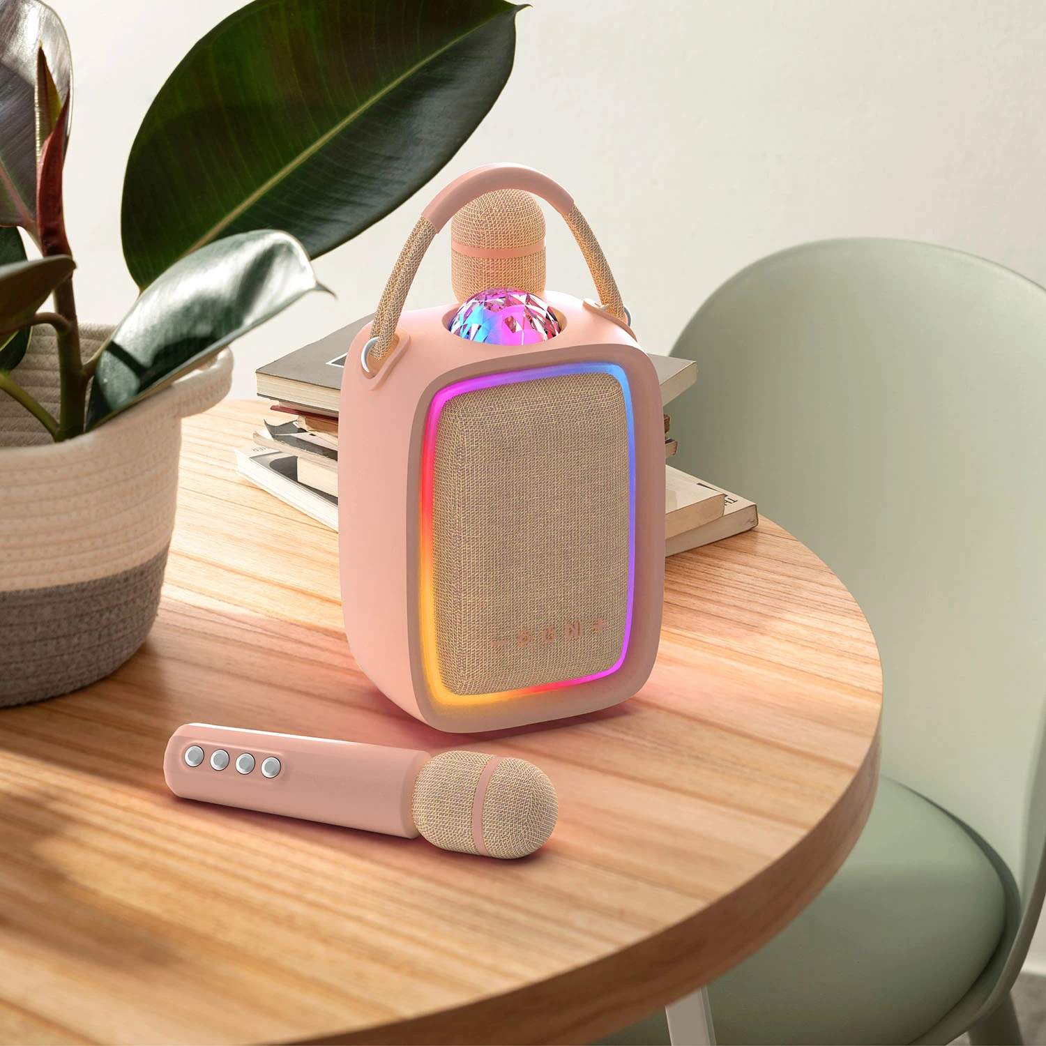 Pink højttaler med discokuglelys og RGB-glød, samt en matchende trådløs mikrofon med fire knapper, på et træbord.