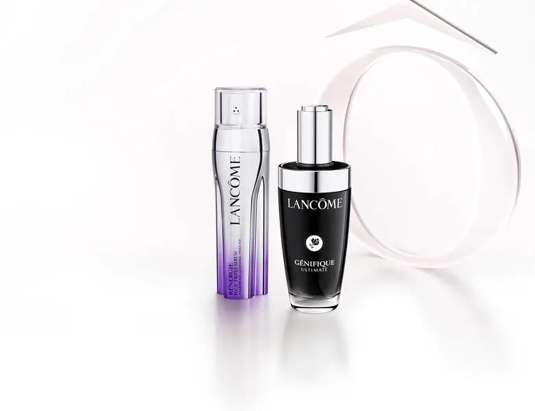 Her ses et billede med to hudplejeprodukter fra Lancôme placeret på en lys, reflekterende overflade mod en lys baggrund. På det ene produkt står “LANCÔME”, og på det andet står “LANCÔME GÉNIFIQUE ULTIMATE”. Bag produkterne ses dekorative, runde elementer.