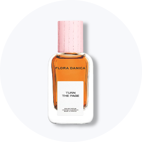 Her ses et billede af en parfumeflakon fra Flora Danica placeret centralt mod en lys, rund baggrund. På flakonen står teksten “FLORA DANICA TURN THE PAGE”. Flakonen er i klart glas med ravfarvet indhold og lyserødt låg.