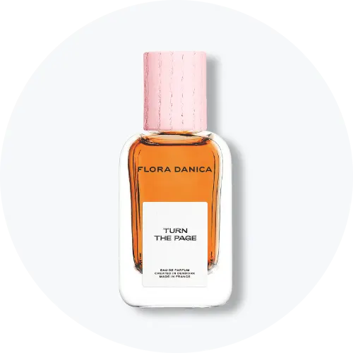 Her ses et billede af en parfumeflakon fra Flora Danica placeret centralt mod en lys, rund baggrund. På flakonen står teksten “FLORA DANICA TURN THE PAGE”. Flakonen er i klart glas med ravfarvet indhold og lyserødt låg.
