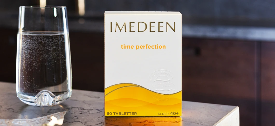 Her ses der et Imedeen produkt i midten ved siden af et glas vand. Baggrunden er grålig som om det står i et køkkenmiljø.
