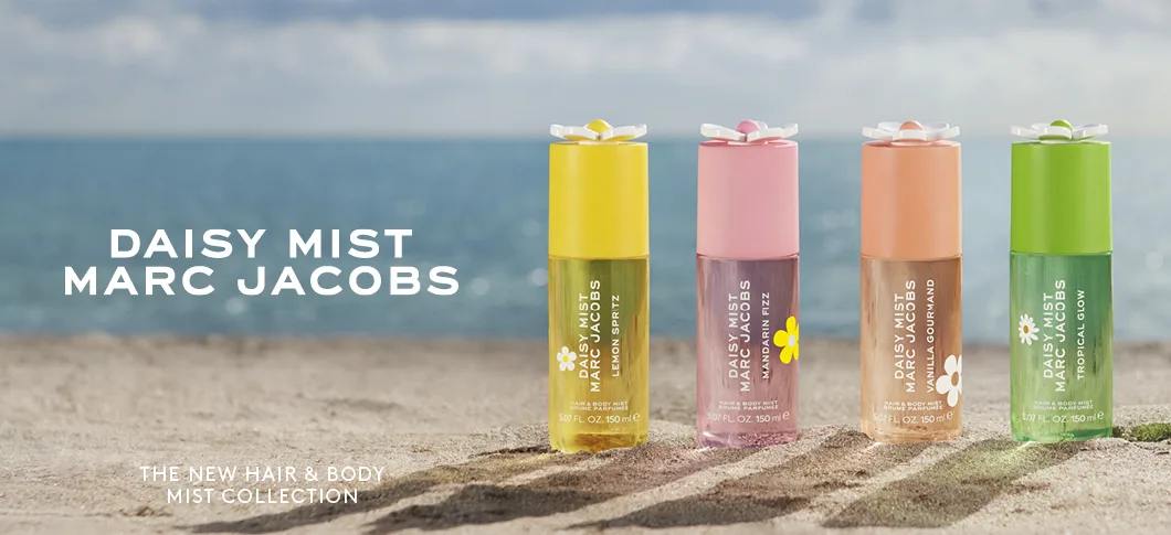 Her ses et billede med fire Daisy Mist Hair & Body Mist-produkter fra Marc Jacobs placeret på sand med havet i baggrunden. Til venstre står teksten “DAISY MIST MARC JACOBS”, og nederst står “THE NEW HAIR & BODY MIST COLLECTION”. Produkterne vises i varianterne Lemon Spritz, Mandarin Fizz, Vanilla Gourmand og Tropical Glow.