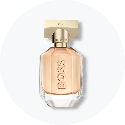 Her ses BOSS The Scent for Her i en transparent parfumeflakon med lyst rosa indhold og guldfarvet låg. BOSS er præget lodret på forsiden af flakonen, som står centralt mod en lys, rund baggrund.