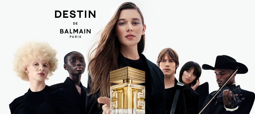 Her ses et billede med flere personer klædt i sort mod en lys baggrund. I midten holder en person en parfumeflakon, og øverst står teksten “DESTIN DE BALMAIN PARIS”. Flakonen er i gyldne nuancer med geometrisk design.