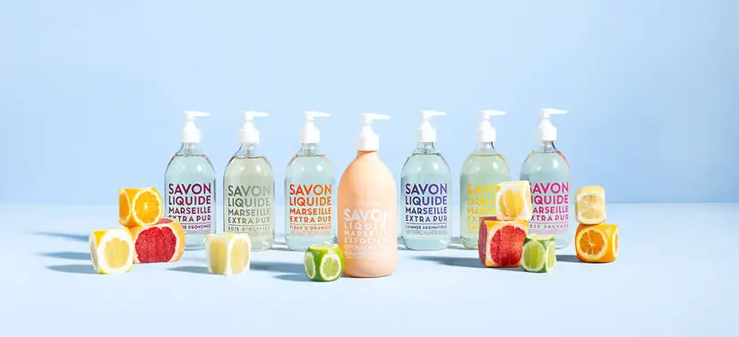 Her ses en lyseblå baggrund med en masse Compagnie de Provence produkter, med tekst i forskellige farver. Der er desuden en masse frugt, som er skåret i kuber.