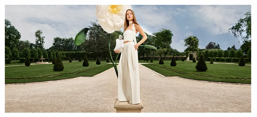 Her ses en kvinde stå i en park foran en stor hvid blomst. Hun holder en Parfums de Marly parfumeflakon.