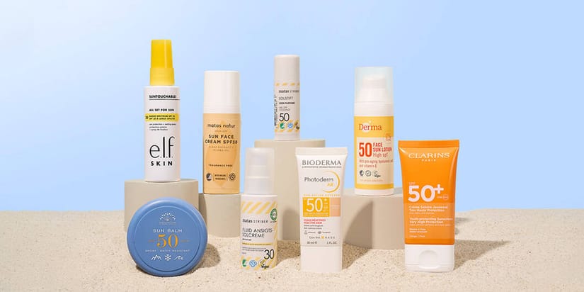 Udvalg af solprodukter i rejsestørrelse fra blandt andet e.l.f. Skin, Matas Natur, Matas Striber, Bioderma, Derma og Clarins står opstillet på sandfarvet underlag mod lyseblå baggrund.