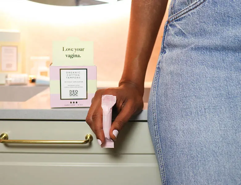 Her ses en kvinde i jeans, som står op af en håndvask. Hun holder en lyserød tampon.