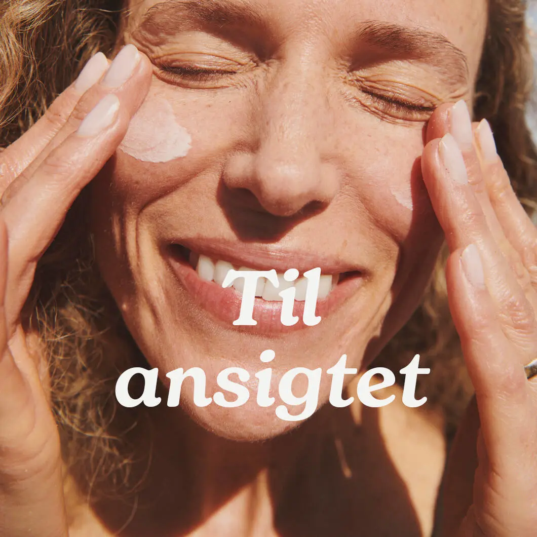 Nærbillede af en smilende kvinde i sollys, der påfører solcreme på begge kinder med fingerspidserne. Midt på billedet står teksten: “Til ansigtet”.