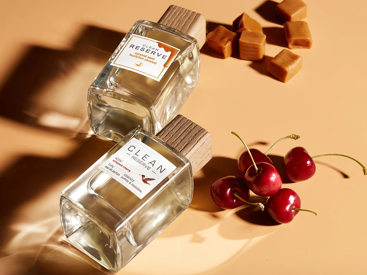 Her ses CLEAN Reserve Caramel Swirl og CLEAN Reserve Whipped Cherry i transparente parfumeflakoner med træfarvede låg. Flakonerne ligger sammen med karameller og røde kirsebær mod en varm beige baggrund.