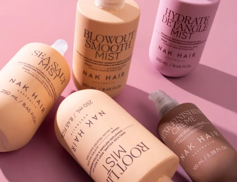 Her ses et billede med stylingprodukter fra NAK Hair placeret mod en lyserød baggrund. Produkterne omfatter blandt andet “Sea Salt Mist”, “Blowout Smooth Mist”, “Hydrate Detangle Mist”, “Root Lift Mist” og “Shine Mist Creme”. Emballagen er i lyserøde, beige og brune nuancer.