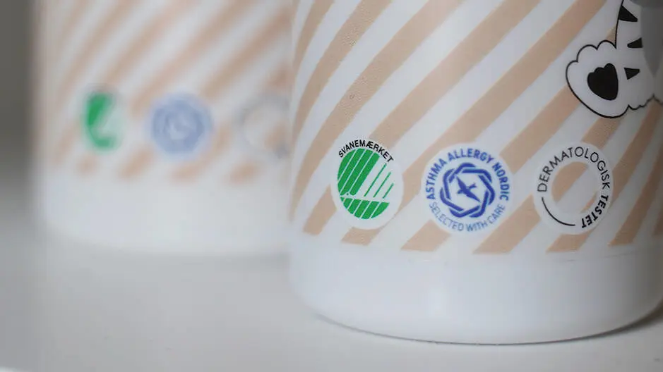 Nærbillede af to lyse emballager med beige og hvide striber. På den forreste emballage ses tre certificeringsmærker: det grønne Svanemærke, Asthma Allergy Nordic-logoet i blå og Dermatologisk Testet-logoet i sort. Baggrunden er sløret, og den anden emballage står ude af fokus i venstre side af billedet.