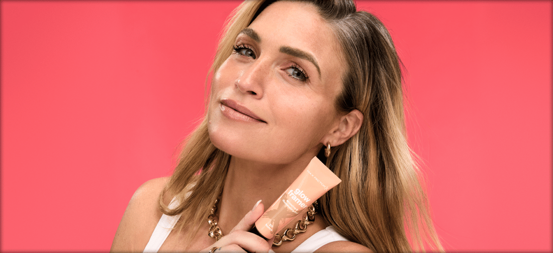 Her ses en rød baggrund med en kvinde som holder et Max Factor "Glow Framer" produkt i hånden. 