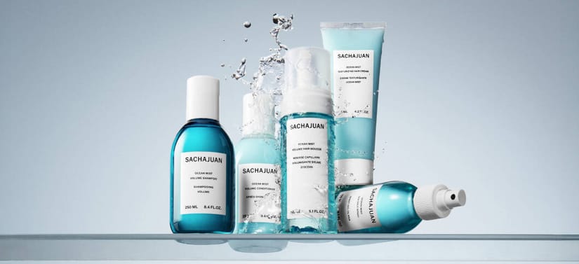 Her ses et billede med flere hårplejeprodukter fra SACHAJUAN placeret samlet på en lys, reflekterende overflade mod en blå baggrund. På produkterne står teksten “SACHAJUAN”. Produkterne er i blå og hvide emballager, og der ses vand, der sprøjter omkring dem.