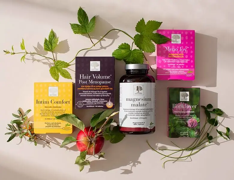 Her ses en lys baggrund med fem forskellige New Nordic produkter, som ligger blandt nogle planter og et æble. 