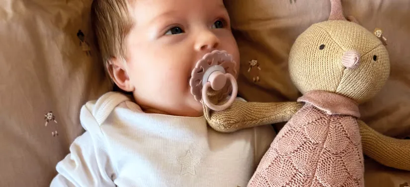 Her ses en baby, som ligger med sin bamse og sut i munden i en seng.
