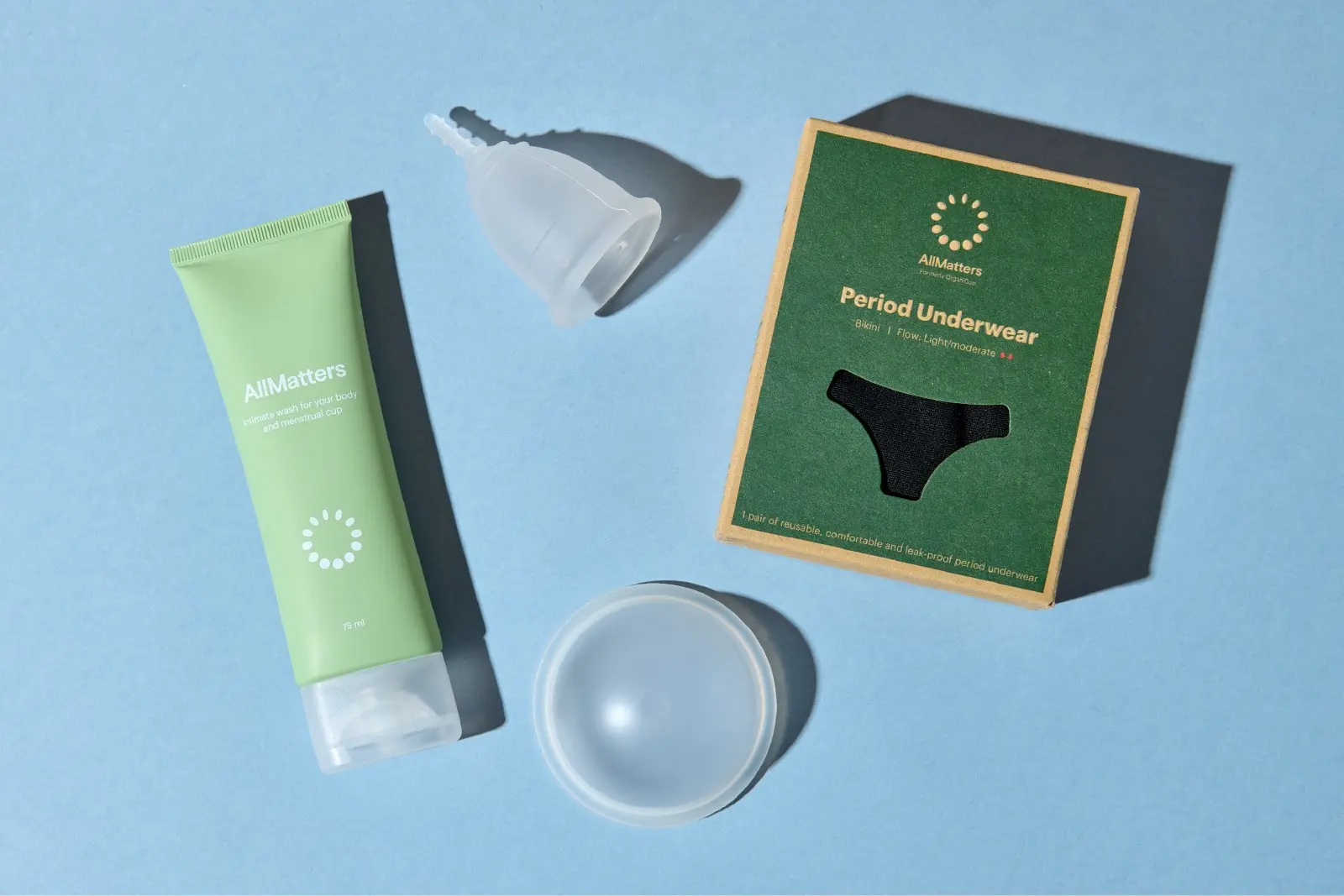 AllMatters produkter til menstruationspleje ligger opstillet på lyseblå baggrund: intimvask i grøn tube, menstruationskop, opbevaringsæske og æske med menstruationstrusser.
