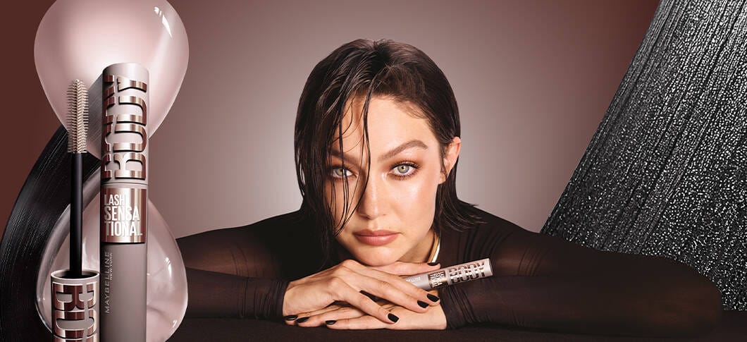 Her ses en kvinde foran en lys lilla baggrund, hvor hun holder en Maybelline Lash Sensational Body Mascara frem. Produktet vises også ved siden af hende til venstre.