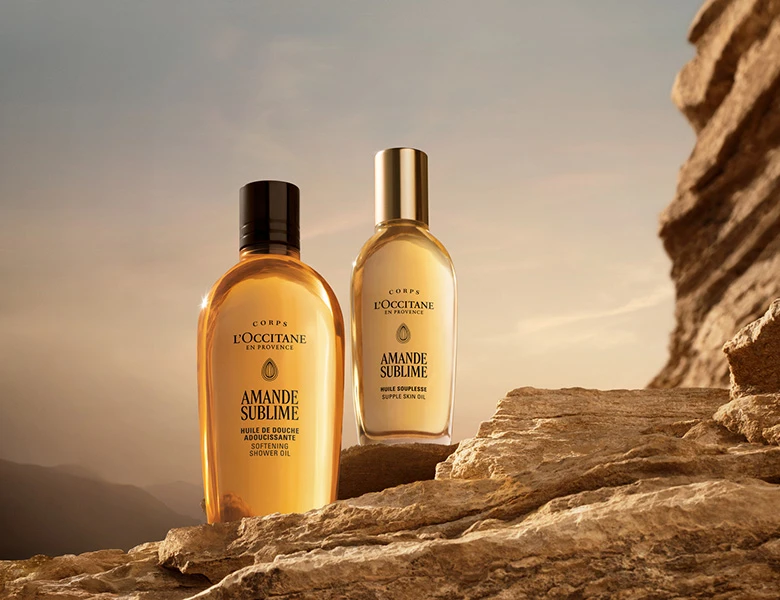 Her ses L’Occitane kropsplejeprodukter i gyldne flasker placeret på klipper i sollys.