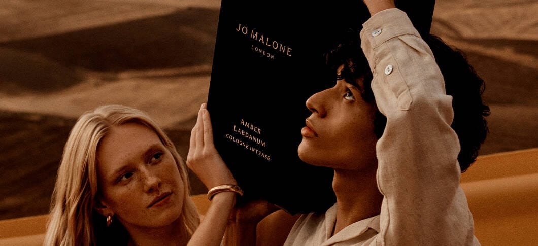 Her ses to personer i noget, der ligner en ørken. Sammen holder de en meget stor Jo Malone parfume.