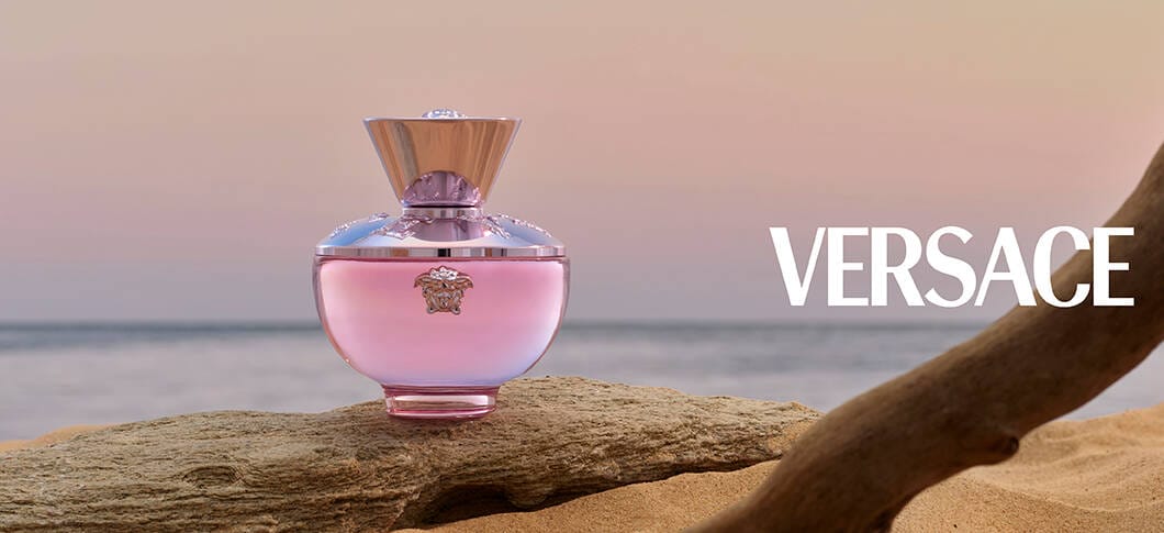 Her ses en Versace Dylan parfume i en pink flakon stående på en sten og med havet som baggund med en lyserød himmel.