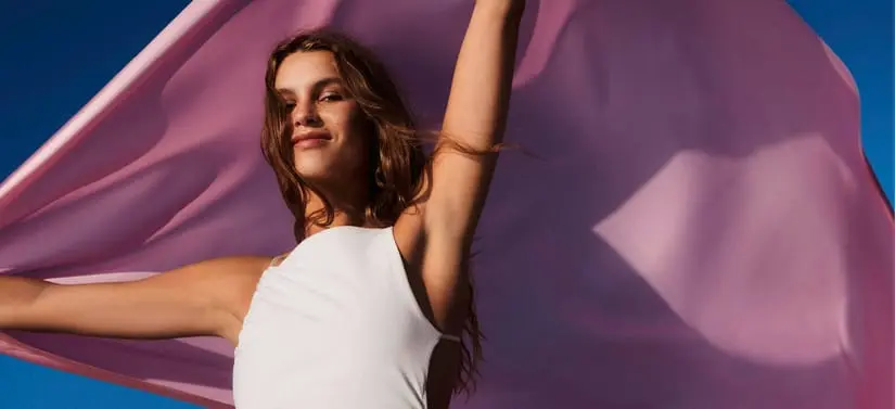 Her ses et billede af en person mod en blå baggrund, hvor en stor pink stofflade holdes op og bevæger sig bag personen. Personen har armene løftet og er iført en hvid top.