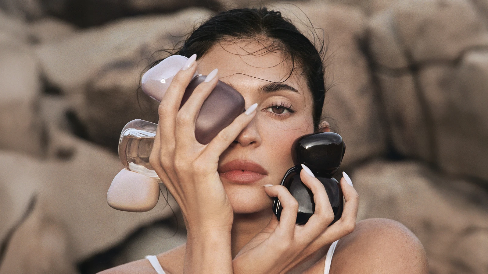 Her ses en person, som holder flere Kylie Cosmetics-parfumeflakoner i lilla, transparente og sorte nuancer omkring ansigtet. Personen har mørkt opsat hår og lyse negle, og billedet er fotograferet mod en neutral baggrund.