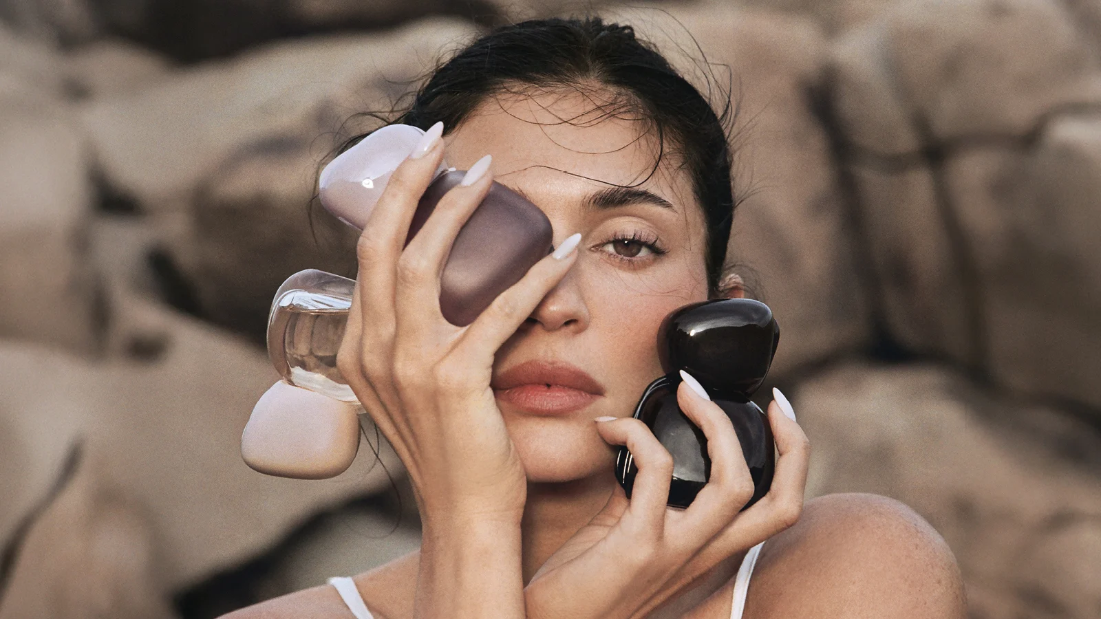 Her ses en person, som holder flere Kylie Cosmetics-parfumeflakoner i lilla, transparente og sorte nuancer omkring ansigtet. Personen har mørkt opsat hår og lyse negle, og billedet er fotograferet mod en neutral baggrund.