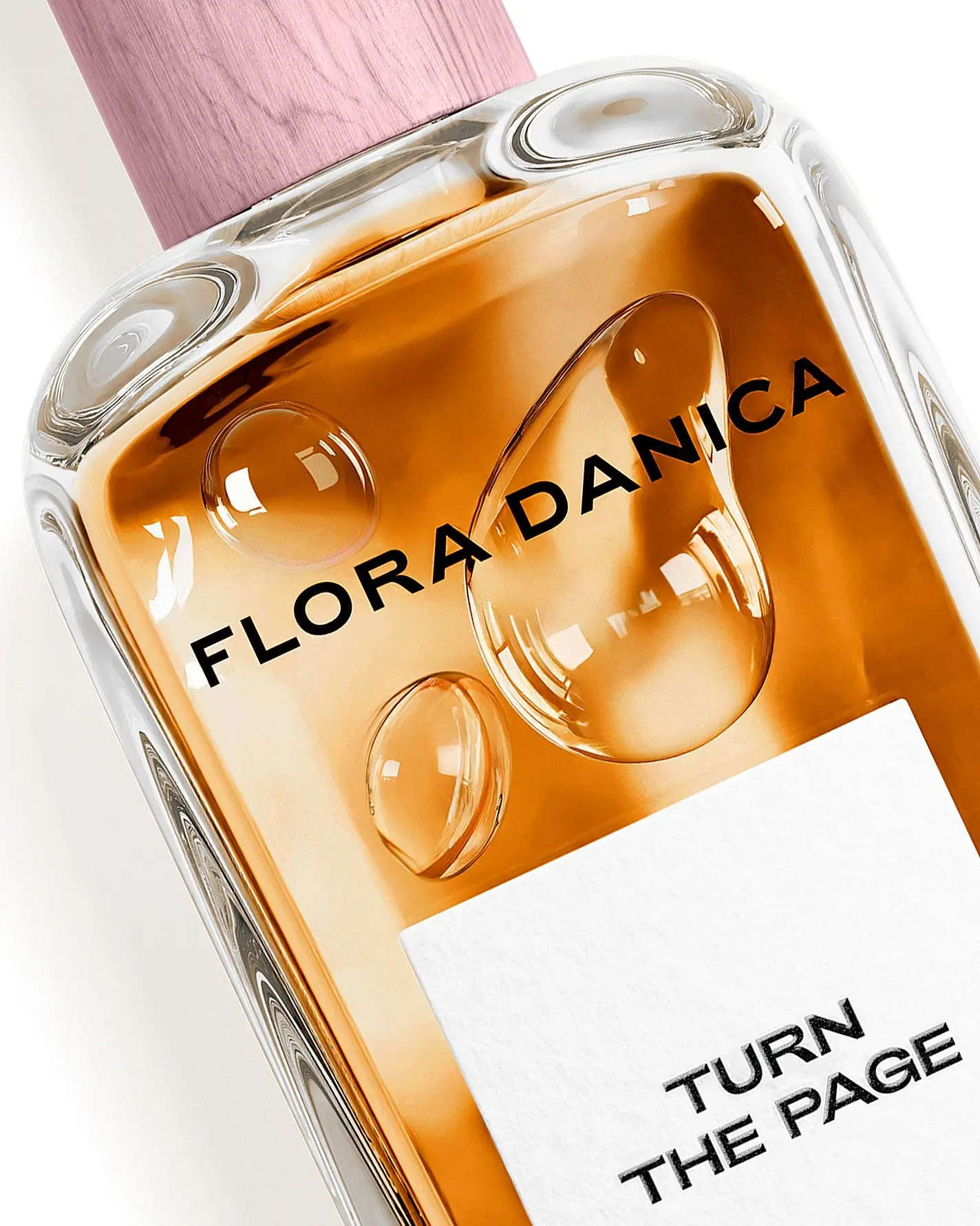 Her ses et nærbillede af en parfumeflakon fra Flora Danica mod en lys baggrund. På flakonen står teksten “FLORA DANICA”. Flakonen har ravfarvet indhold og et lyserødt låg.