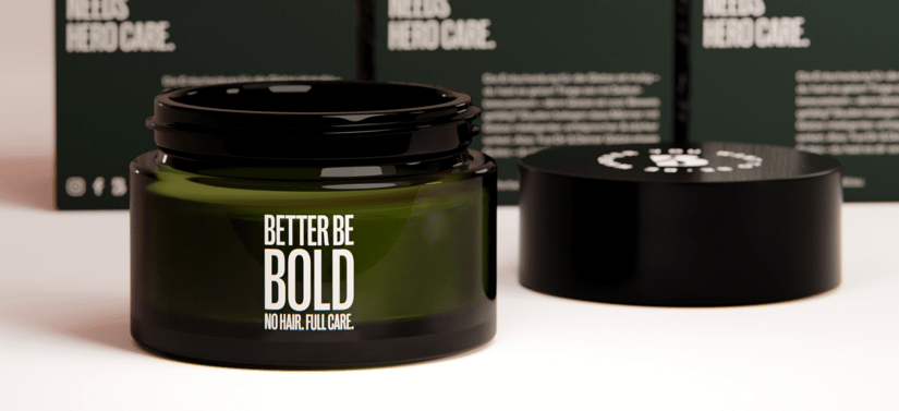 Åben mørkegrøn Better Be Bold krukke med sort låg står på lys overflade, mens produktæsker står sløret i baggrunden.