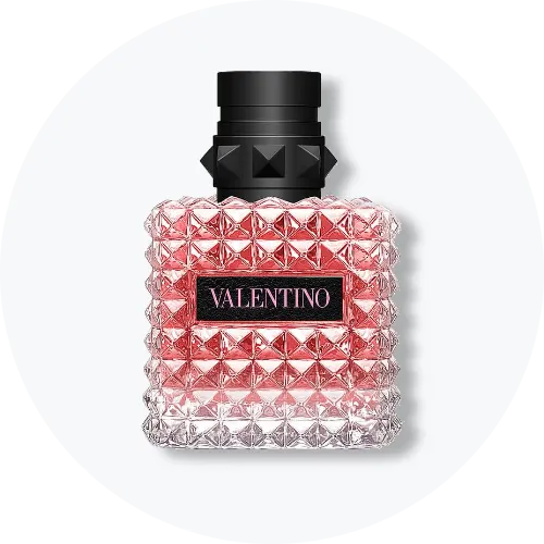 Her ses en Valentino-parfumeflakon med rosa indhold og et facetteret design med karakteristiske nittedetaljer. Flakonen har sort låg og et sort Valentino-logo på forsiden og står centralt mod en lys, rund baggrund.
