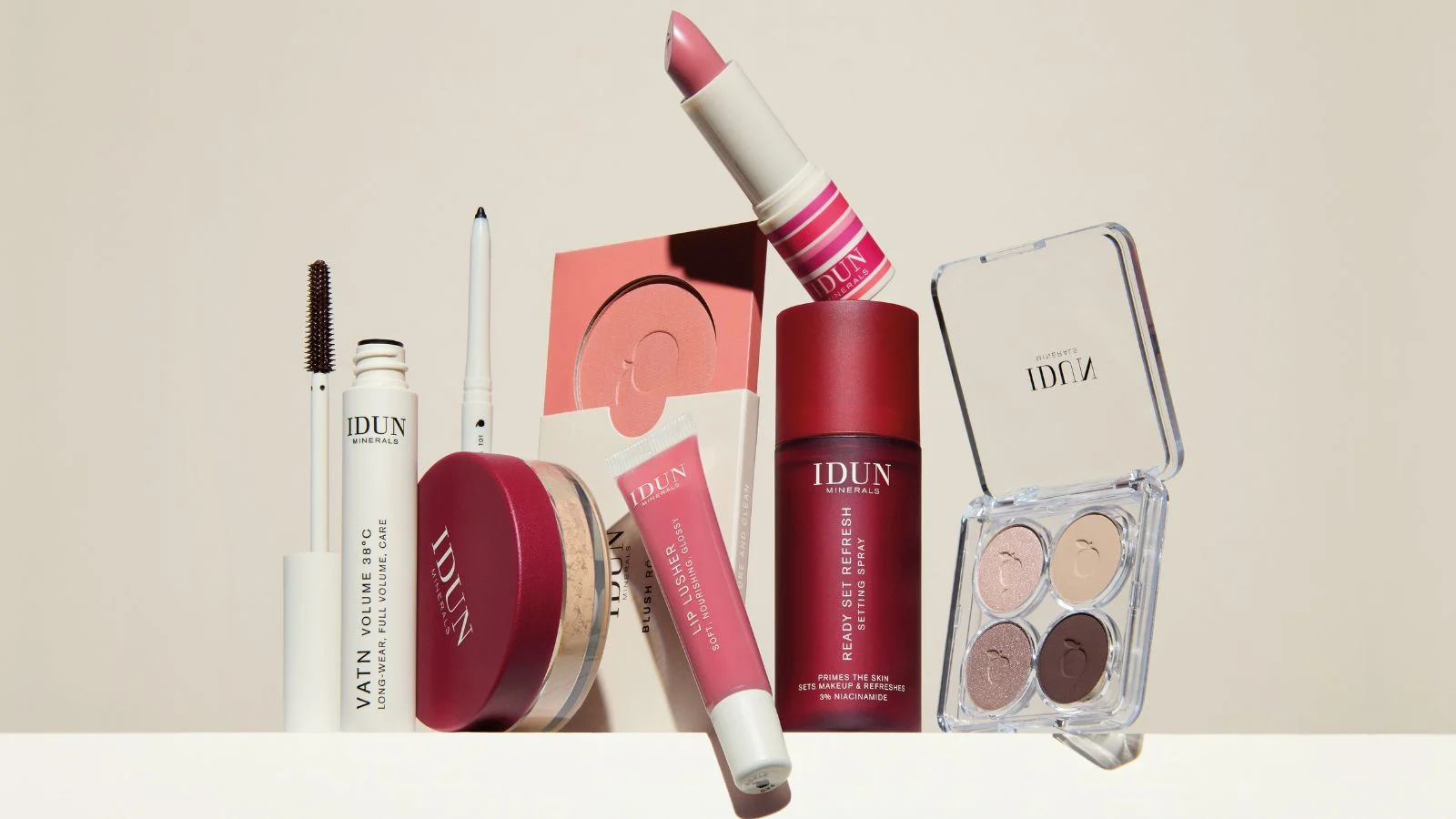 Her ses et billede med et udvalg af makeup fra IDUN Minerals arrangeret mod en lys baggrund. Produkterne omfatter blandt andet Vatn Volume 38°C Mascara, en eyeliner, en læbestift, Lip Lusher lipgloss, en blush, løspudder, Ready Set Refresh Setting Spray og en åben øjenskyggepalette med fire nuancer. Produkterne er opstillet samlet for at fremhæve forskellige kategorier inden for makeup, herunder øjne, læber, ansigt og setting.