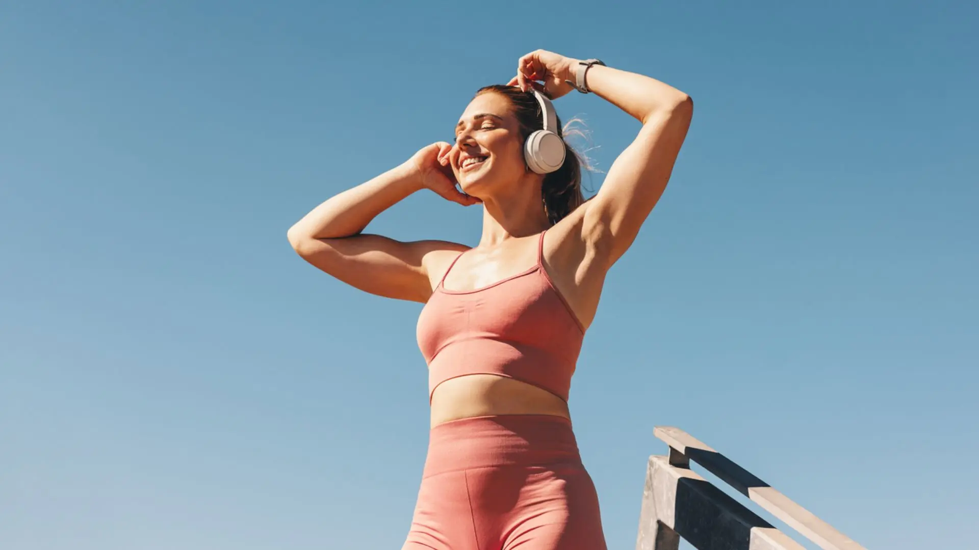En smilende kvinde i lyserødt sportstøj lytter til musik med hvide hovedtelefoner under en klar blå himmel.