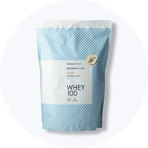 En pose med Matas Striber Whey 100 proteinpulver står på en lys grå, cirkulær baggrund. På posen ses et mærke med teksten "Vanilje".