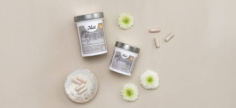 To lyse Nani bøtter med præ- og probiotika ligger på lys træbaggrund omgivet af hvide blomster, kapsler og en lille skål med kapsler.