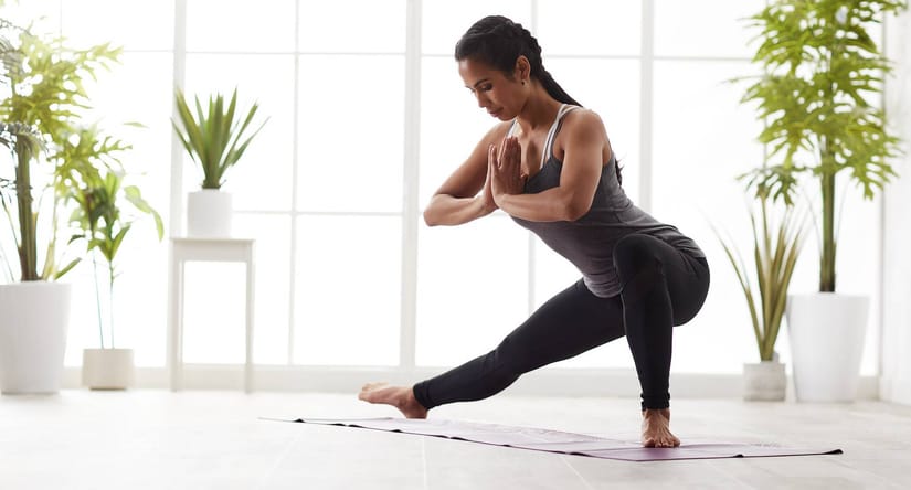 Kvinde i træningstøj laver en yogaøvelse på en yogamåtte i et lyst rum med store vinduer og grønne planter i baggrunden.