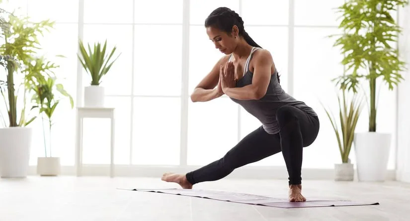 Kvinde i træningstøj laver en yogaøvelse på en yogamåtte i et lyst rum med store vinduer og grønne planter i baggrunden.