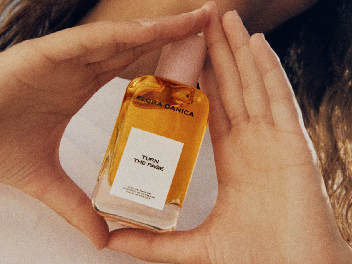Her ses et nærbillede af en person, der holder en parfumeflakon mellem hænderne. Parfumen er fra Flora Danica, og på flasken står teksten “TURN THE PAGE”. Flakonen indeholder en gylden væske og har et lyserødt låg.