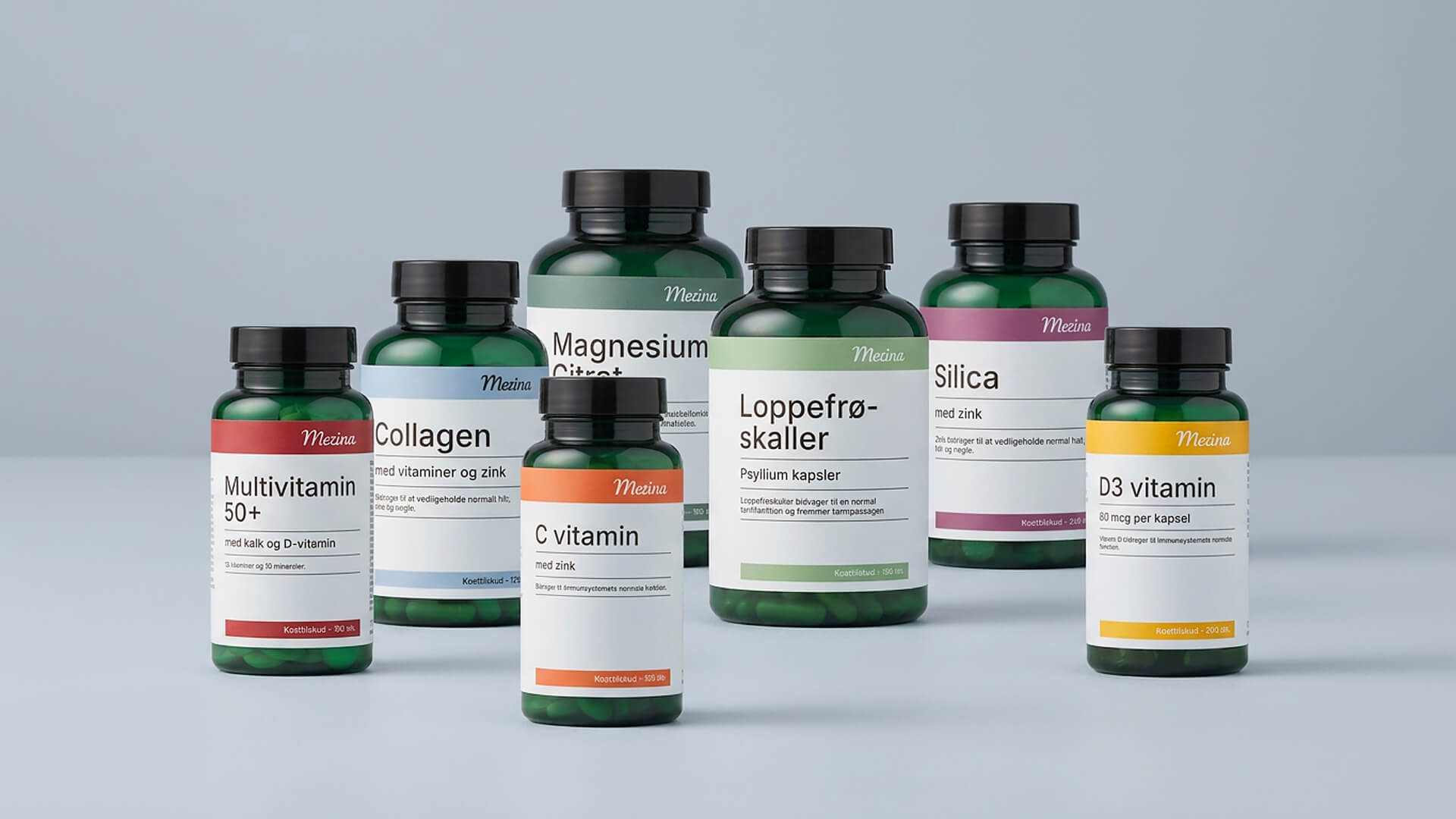 Seks grønne Mezina kosttilskudsbøtter står samlet på lyseblå baggrund, blandt andet multivitamin 50+, collagen, C vitamin, magnesium citrat, loppefrøskaller, silica og D3 vitamin.