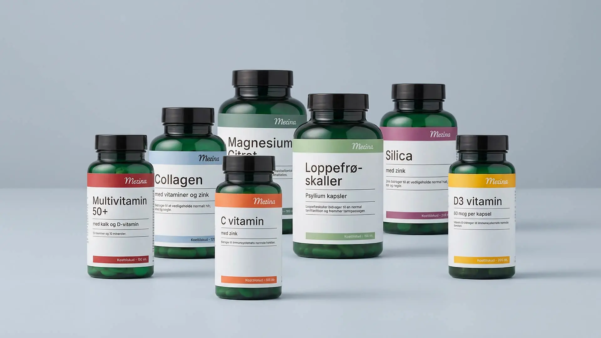 Seks grønne Mezina kosttilskudsbøtter står samlet på lyseblå baggrund, blandt andet multivitamin 50+, collagen, C vitamin, magnesium citrat, loppefrøskaller, silica og D3 vitamin.